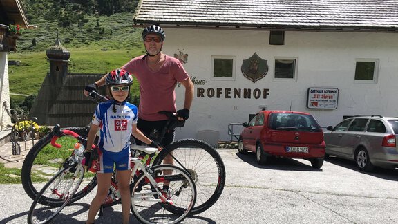 A cyclist and a child stand with their bicycles in front of a building with the inscription "Rofenhof". The surroundings are green and mountainous. - Haus Albin - Sölden
