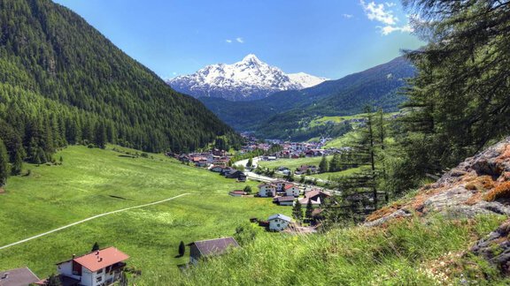 A picturesque mountain landscape with snow-capped peaks and a green meadow. In the valley lies a small village, surrounded by trees and mountains. - Haus Albin - Sölden