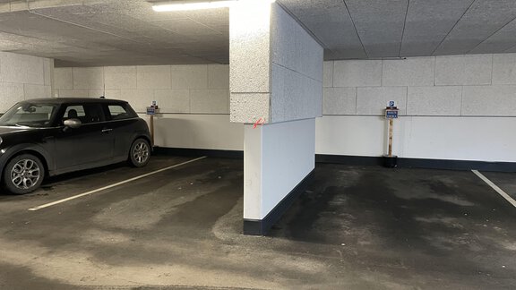 An empty parking space in an underground garage with a black car on the left side. The walls are white, and the floor is dirty. - HAUS ERIKA apartments - Sölden