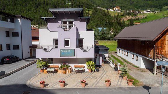 A modern building in purple with a well-maintained entrance area and flower pots. In the background, green mountains can be seen. - Alpenapart Saphir - Sölden