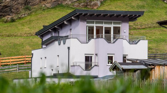 A modern, multi-storey house in a rural setting. It has large windows and a balcony overlooking the green meadow. - Alpenapart Saphir - Sölden
