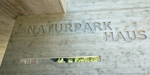 Ein modernes Gebäude mit der Aufschrift „NATURPARK HAUS“ auf einer Betonwand. Es gibt ein Fenster, das Ausblicke auf die Natur ermöglicht. - Naturpark Ötztal - Naturpark Haus - Längenfeld