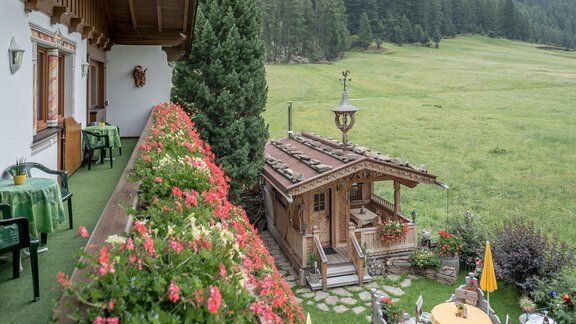 Ein gemütlicher Balkon mit bunten Blumen und Blick auf eine grüne Wiese. Im Hintergrund steht ein rustikales Holzhaus, umgeben von Bäumen und Berglandschaft. - Apart Wiesengrund - Sölden
