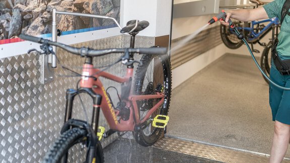 Ein Fahrrad wird an einem Waschplatz gereinigt. Der Benutzer sprüht Wasser auf das Rad, um Schmutz zu entfernen. - Garni Ötztal ROOMS & APARTS - Zwieselstein