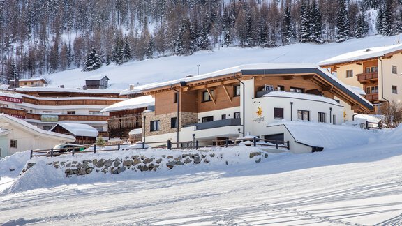 A picturesque winter landscape with snow-covered hills and a modern alpine lodge. In the background, traditional buildings are visible in the tranquil surroundings. - Apart Sonnhof - Sölden