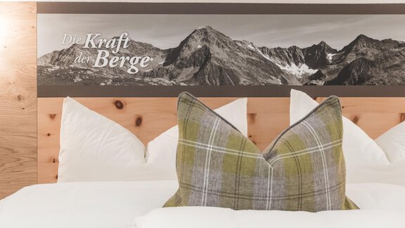 Ein modernes Schlafzimmer mit einem gemütlichen Kissen und einer Wandverkleidung, die beeindruckende Berge zeigt. Der Schriftzug „Die Kraft der Berge“ betont die alpine Atmosphäre. - Garni Ötztal ROOMS & APARTS - Zwieselstein