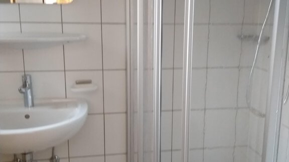 A small bathroom with a shower, a sink, and a mirror. The walls are tiled with white tiles. - Michls Hoamat Appartement - Sölden