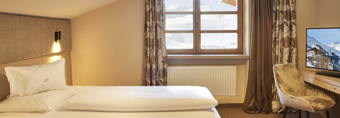 Hotel-Bergwelt-Obergurgl-Einzelzimmer-B1 | © Art & Relax Hotel Bergwelt (CC BY) A modern hotel room with a large window that lets in plenty of natural light. It features a cozy bed, a desk, and a television. - Art & Relax Hotel Bergwelt - Obergurgl