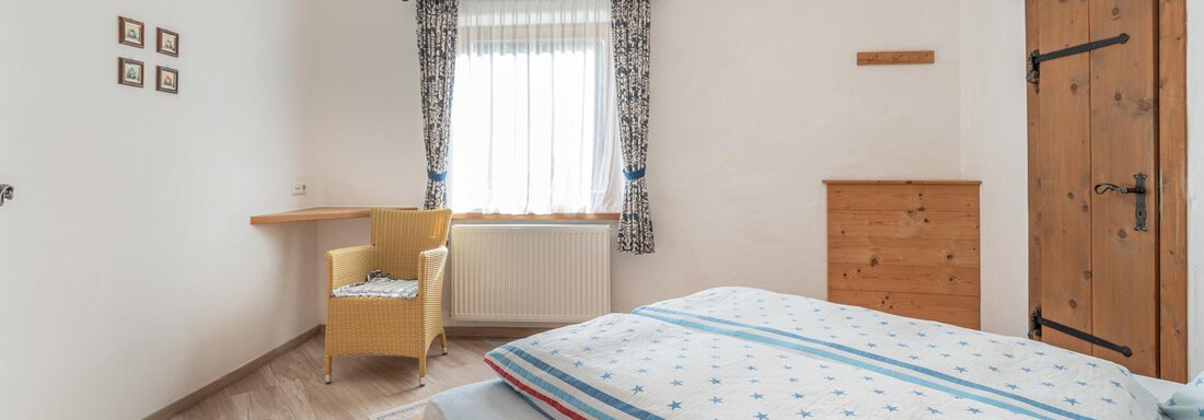 Ein helles Schlafzimmer mit einem Doppelbett und blauen Dekorationen. Es gibt einen Arbeitstisch, einen Stuhl und einen Holzschrank. - Ferienwohnung Alt Poschach - Obergurgl 