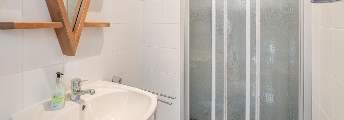 Ein modernes Badezimmer mit einer Dusche und einem Waschbecken. Die Wände sind weiß gefliest und ein Holzmöbel mit Spiegel ist vorhanden. - Ferienwohnung Alt Poschach - Obergurgl 