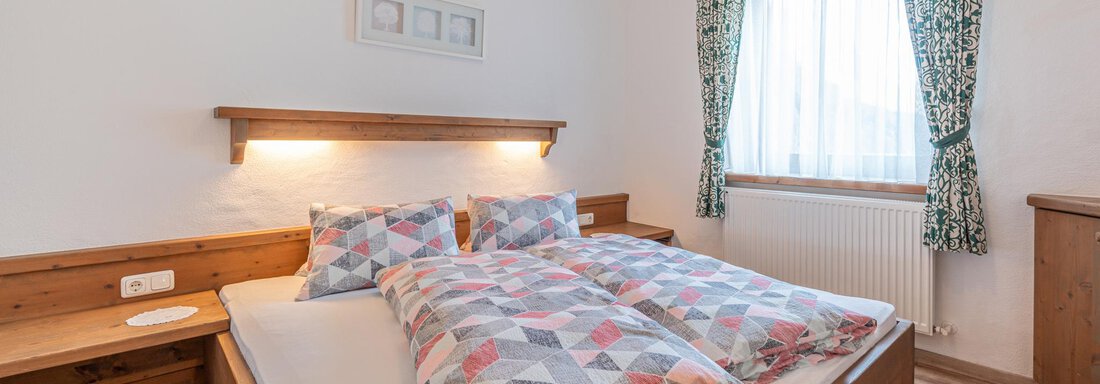 Ein gemütliches Schlafzimmer mit einem Doppelbett und bunten Bettdecken. Das Fenster lässt viel Licht herein und die Einrichtung ist aus Holz. - Ferienwohnung Alt Poschach - Obergurgl 