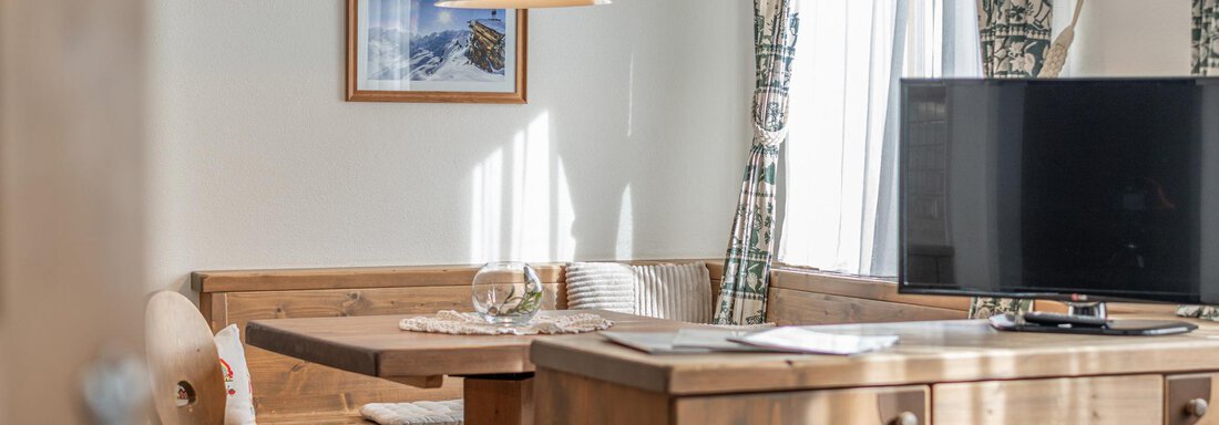 Ein gemütlicher Wohnbereich mit Holzmöbeln und einem Esstisch. Helle Fenster lassen viel Tageslicht herein, und ein Fernseher ist sichtbar. - Ferienwohnung Alt Poschach - Obergurgl 