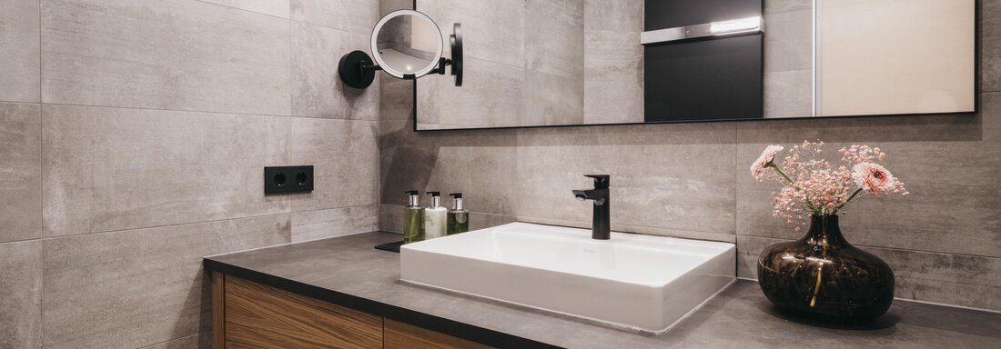 A modern bathroom with a large mirror and an elegant sink. The walls are painted in neutral tones, and a beautiful bouquet adds a splash of color. - The Crystal VAYA Unique - Obergurgl 
