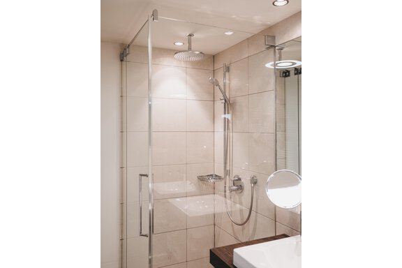 A modern shower cabin with clear glass walls and elegant tiles. In the background, a sink and a lit mirror can be seen. - The Crystal VAYA Unique - Obergurgl 
