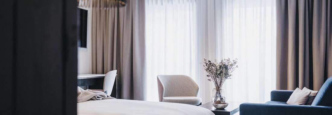VAYA The Crystal A modern room with a large window and soft curtains. There is a cozy armchair, a couch, and a vase with flowers. - The Crystal VAYA Unique - Obergurgl