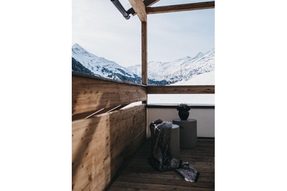 A balcony with wooden cladding and a view of snow-capped mountains. A blanket and a small table complete the cozy atmosphere. - The Crystal VAYA Unique - Obergurgl 