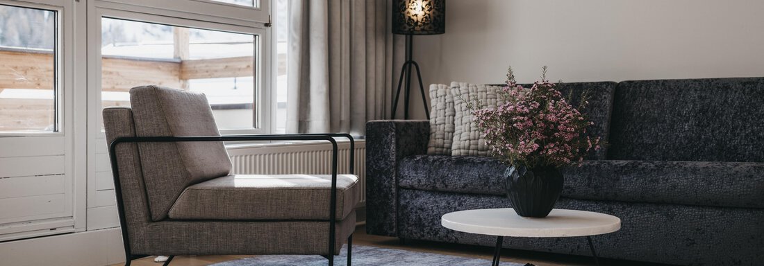 A modern living room with a gray armchair and a dark couch. A round table with a flower vase is placed on a light carpet. - The Crystal VAYA Unique - Obergurgl 