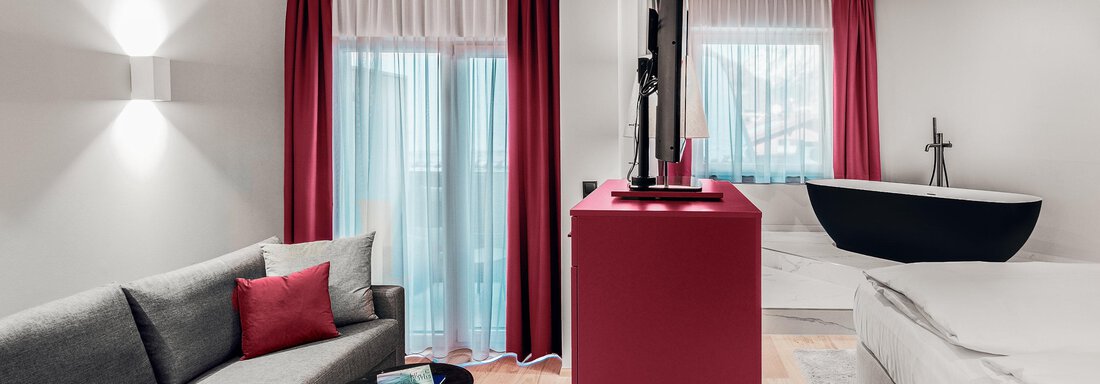 Ein modernes Hotelzimmer mit einem grauen Sofa, einem roten Fernsehtisch und großen Fenstern. Im Hintergrund sieht man ein modernes Bett und eine freistehende Badewanne. - AlpenParks The Secret Sölden - Sölden