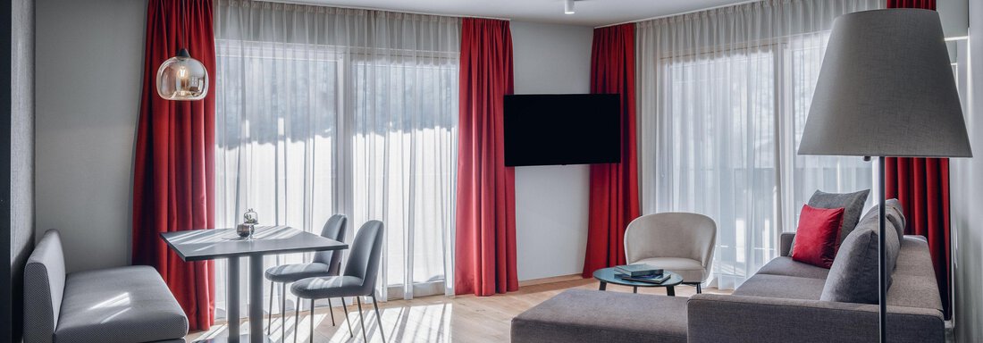 Ein modernes Wohnzimmer mit großen Fenstern und rotem Vorhang. Die Einrichtung besteht aus einem bequemen Sofa, einem Tisch und Stühlen. - AlpenParks The Secret Sölden - Sölden