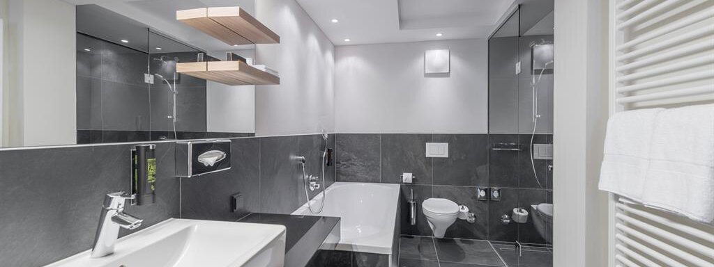 Ein modernes Badezimmer mit eleganten, schwarzen Fliesen und einer großen Badewanne. Helle Beleuchtung und spiegelnde Oberflächen schaffen eine angenehme Atmosphäre. - AlpenParks The Secret Sölden - Sölden