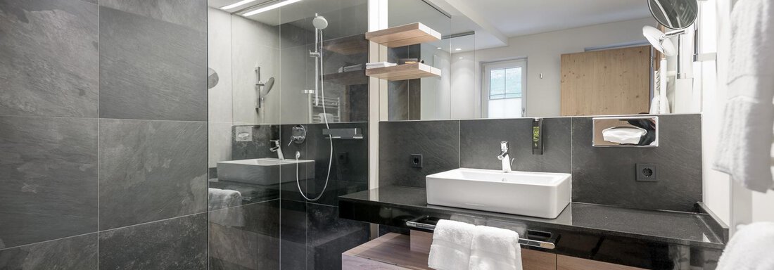 Ein modernes Badezimmer mit schwarzen Fliesen und einer eleganten Dusche. Der Waschtisch aus Holz und die großen Spiegel schaffen ein stilvolles Ambiente. - AlpenParks The Secret Sölden - Sölden