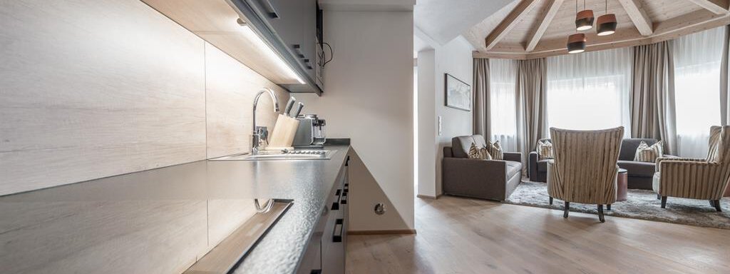 Eine moderne Küche mit einem eleganten Design und Holzoberflächen. Im Hintergrund sieht man ein komfortables Wohnzimmer mit gemütlichen Sitzgelegenheiten und großen Fenstern. - AlpenParks The Secret Sölden - Sölden