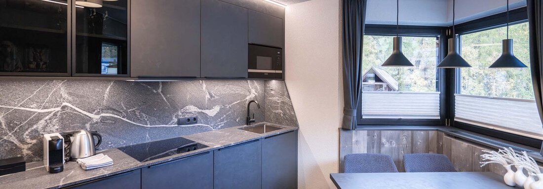 Eine moderne Küche mit dunklen Schränken und elegenen Marmorelementen. Am Tisch sind zwei Stühle und große Fenster sorgen für viel Tageslicht. - Apart Reinstadler - Sölden