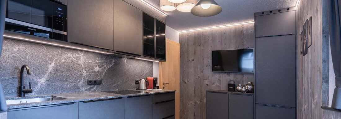 Eine moderne Küche mit eleganten, schwarzen Schränken und einer hellen, stilvollen Beleuchtung. Der Essbereich besteht aus einem einfachen Tisch und gemütlichen Stühlen. - Apart Reinstadler - Sölden