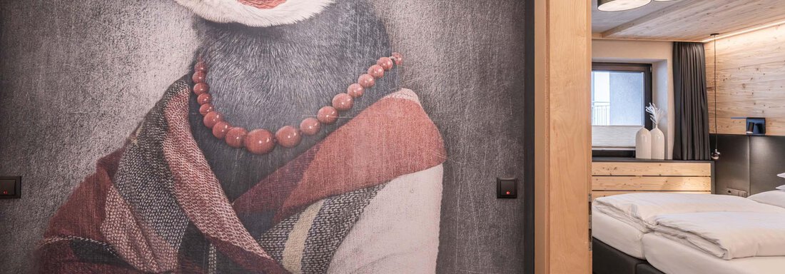 Ein kreatives Wandbild eines Penguins in eleganter Kleidung und mit einer Perlenkette. Im Hintergrund ist ein modernes Zimmer mit einem Doppelbett und einem gemütlichen Ambiente sichtbar. - Apart Reinstadler - Sölden