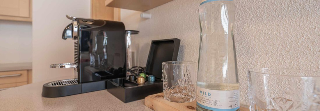 Eine moderne Küchenecke mit einer Espresso-Maschine und einer Glasflasche Wasser. Neben der Flasche stehen zwei Gläser auf einem Holzbrett. - Apart Reinstadler - Sölden