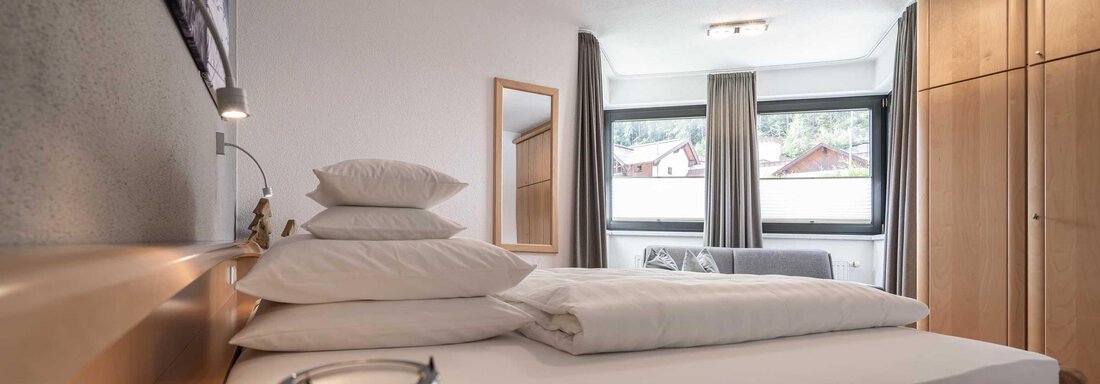 Ein modernes Schlafzimmer mit einem großen Bett und vielen Kissen. Große Fenster sorgen für viel Tageslicht. - Apart Reinstadler - Sölden