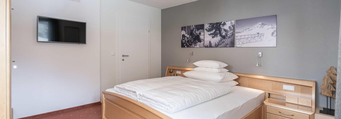 Ein gemütliches Schlafzimmer mit einem Doppelbett und modernem Holzdesign. An der Wand hängen Bilder von schneebedeckten Bergen, und ein Fernseher ist an der Wand montiert. - Apart Reinstadler - Sölden