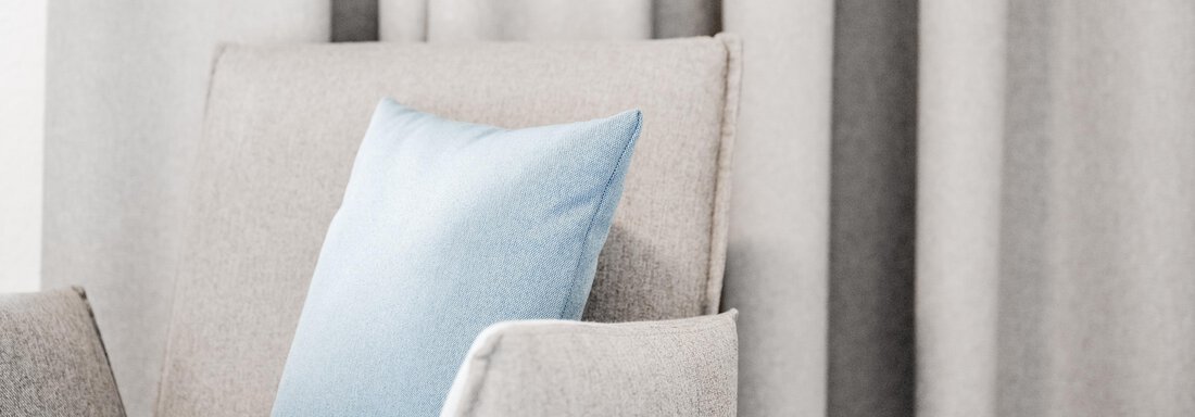 Ein gemütlicher Sessel mit einem blauen Kissen steht vor einem neutralen Vorhang. Die Farben sind weich und laden zum Entspannen ein. - Appartement Montanara - Sölden