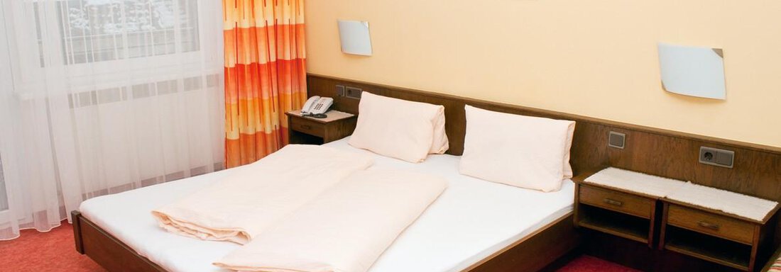 Ein einfaches Hotelzimmer mit einem Doppelbett und orangefarbenen Vorhängen. An der Wand hängen zwei Bilder, und es gibt einen Schreibtisch sowie ein Telefon. - Appartement Viertler - Sölden