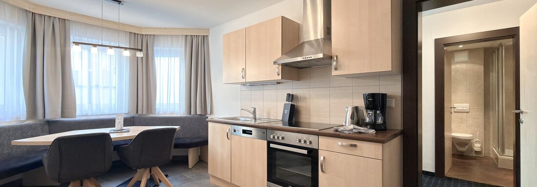 A modern kitchen with light wooden cabinets and a cozy seating area. The light pours in through large windows, creating an inviting atmosphere. - Appartementhaus A CASA "Kristall" - Sölden