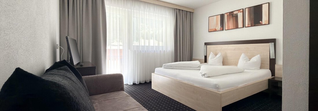 A modern hotel room with a large bed and a sofa. The windows provide plenty of daylight. - Appartementhaus A CASA "Kristall" - Sölden