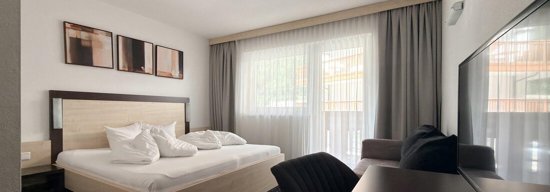 A modern bedroom with a double bed and white pillows. The room is bright and has large windows with curtains. - Appartementhaus A CASA "Kristall" - Sölden