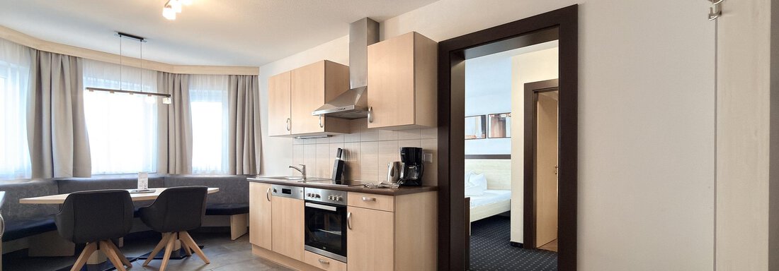 Kitchen in a modern living space with wooden cabinets and a cooking area. The room is bright and has large windows with curtains. - Appartementhaus A CASA "Kristall" - Sölden