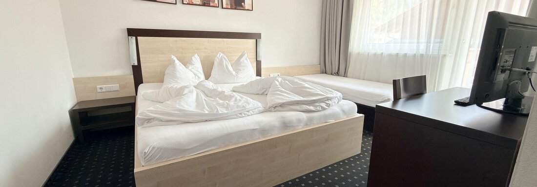 A modern bedroom with a large bed and two pillows. The room has a desk, a television, and plenty of daylight through the windows. - Appartementhaus A CASA "Kristall" - Sölden