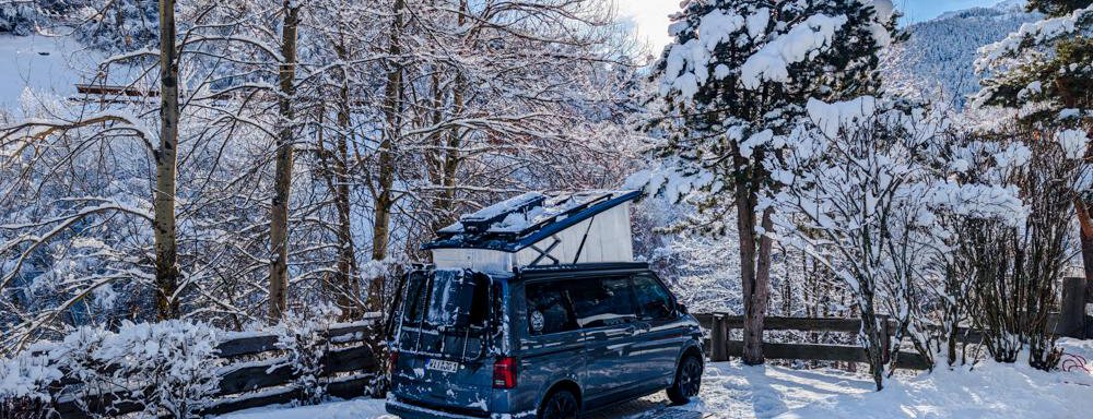 Stellplatz Deluxe A blue campervan is parked in a snow-covered landscape. Tall trees and a clear sky complement the wintry scene. - Camping Sölden - Sölden