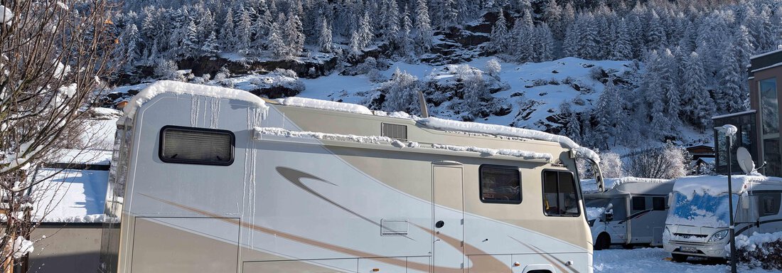 A camper stands in a snowy landscape. In the background, snow-covered mountains and trees can be seen. - Camping Sölden - Sölden