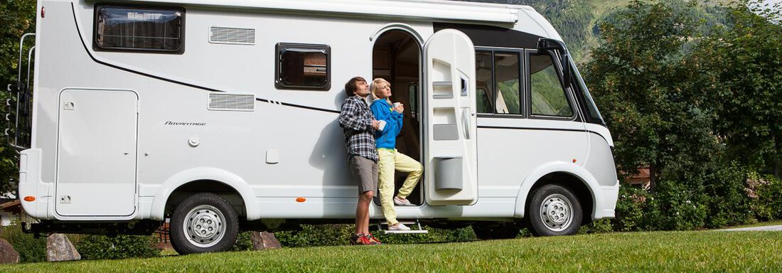 A camper van is parked on a green meadow with mountains in the background. Two people are standing in front of it and enjoying the view. - Camping Sölden - Sölden