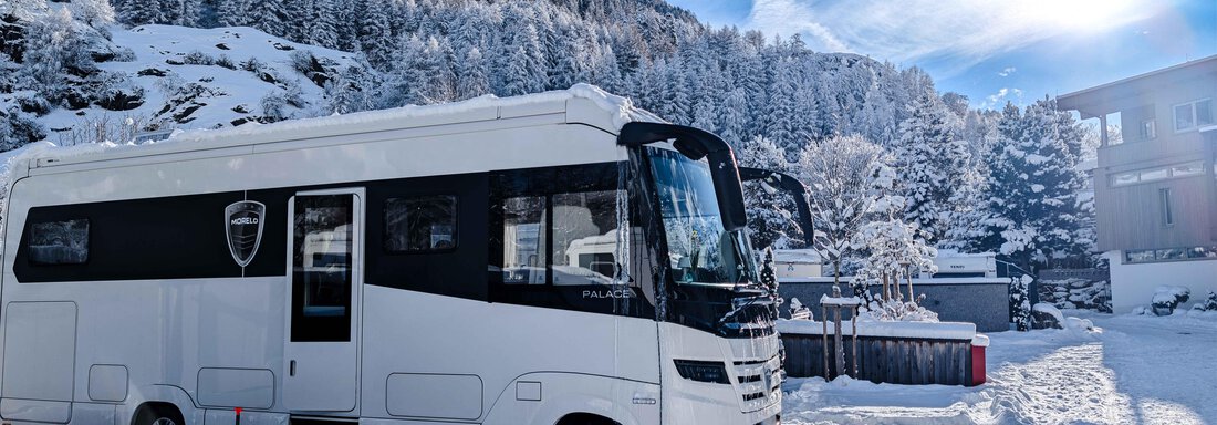 CS_Platz-Winter-komfort A motorhome is situated in a snowy environment under a blue sky. In the background, snow-covered trees and hills can be seen. - Camping Sölden - Sölden