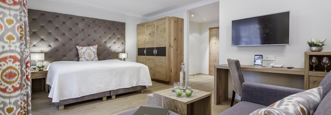 Ein modernes Hotelzimmer mit einem gemütlichen Bett und einem grauen Sofa. Die Einrichtung ist hell und einladend, mit einem Fernseher und einem kleinen Tisch. - Das Central - Alpine . Luxury . Life - Sölden