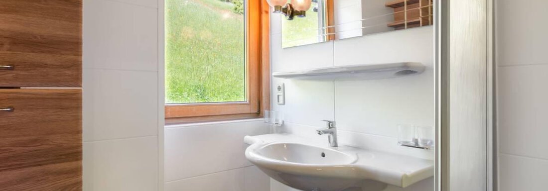 Ein modernes Badezimmer mit einem Waschbecken und einem großen Spiegel. Durch das Fenster fällt natürliches Licht und zeigt eine grüne Landschaft. - Garni Nadine - Sölden