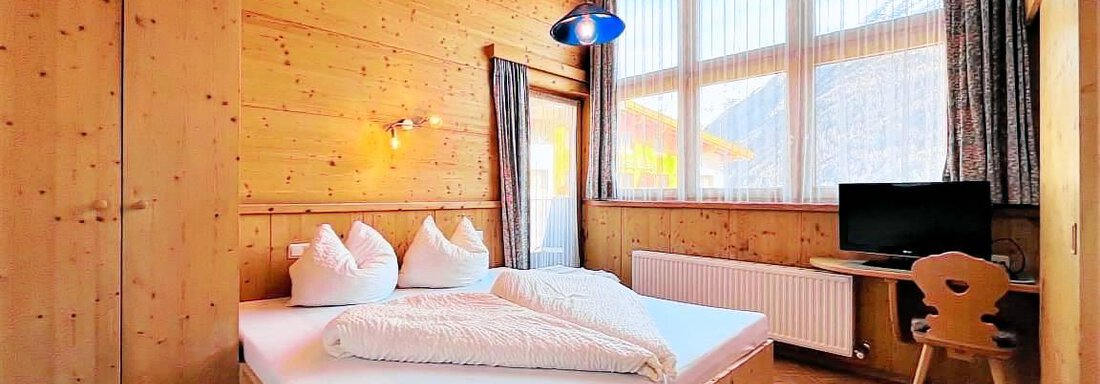 Ein gemütliches Schlafzimmer aus Holz mit einem großen Fenster und natürlichem Licht. Es gibt ein Doppelbett und einen Schreibtisch mit Stuhl. - Garni Nadine - Sölden