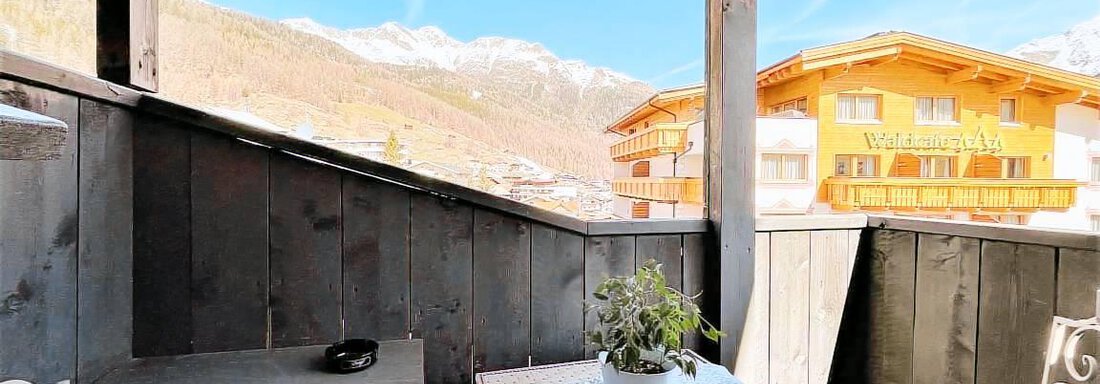 Ein gemütlicher Balkon mit einem Tisch und Stühlen, umgeben von Holzverkleidung. Im Hintergrund sind Berge und eine helle blaue Himmel zu sehen. - Garni Nadine - Sölden