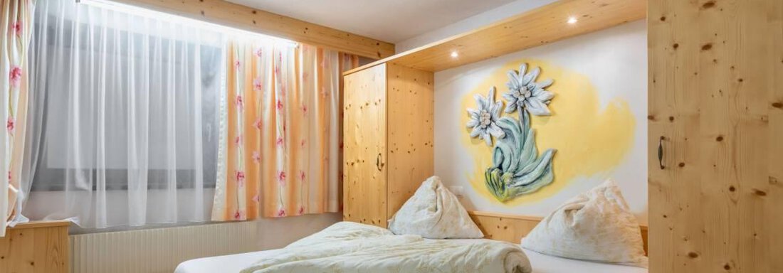 Ein gemütliches Schlafzimmer mit Holzmöbeln und einem großen Fenster. An der Wand befindet sich eine schöne Blumenmalerei. - Garni Nadine - Sölden