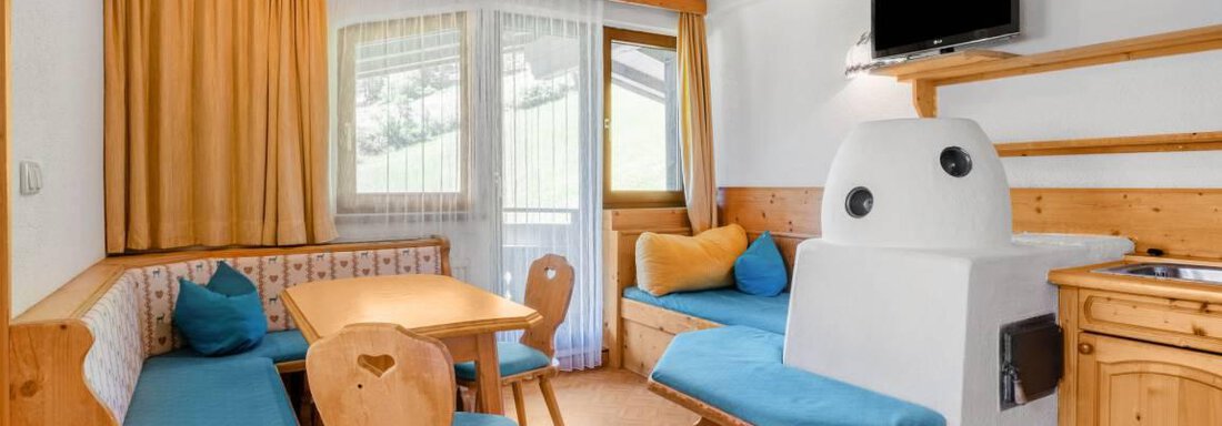 Ein gemütlicher Wohnbereich mit Holzverkleidung und blauen Sitzen. Ein Tisch steht zentral im Raum, und große Fenster bieten Tageslicht. - Garni Nadine - Sölden