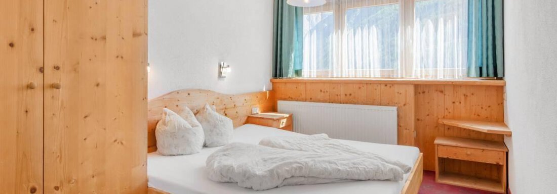 Ein gemütliches Schlafzimmer mit Holzmöbeln und einem großen Fenster. Das Zimmer ist hell und freundlich mit einem Doppelbett und einem kleinen Tisch. - Garni Nadine - Sölden